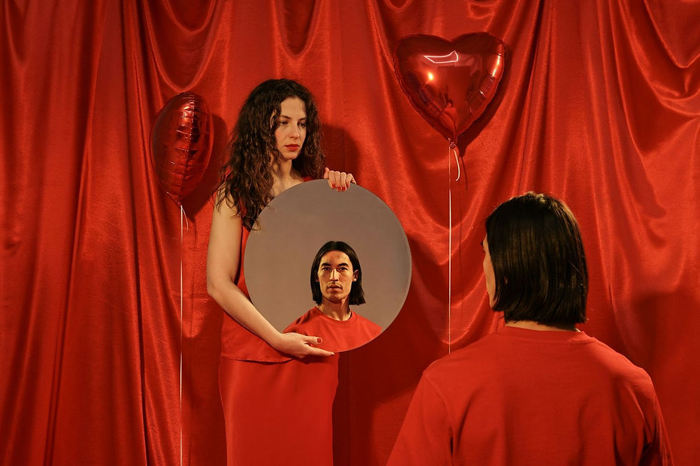 A woman in red holds a mirror reflecting a man's face. Red curtains and two heart-shaped balloons create a dramatic setting.