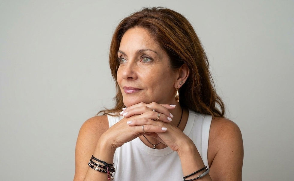 Woman in a white sleeveless top rests her chin on clasped hands, gazing aside against a plain gray background.