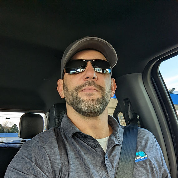 Man in a car wearing a cap and sunglasses, seatbelt on, looking ahead with a neutral expression.