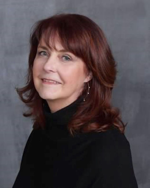 Woman with auburn hair and a black turtleneck smiles gently against a gray background. Mood is calm and friendly.