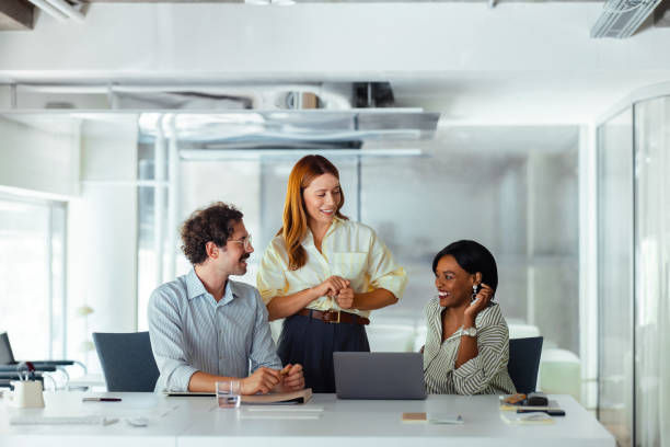 Three people in an office collaborate around a laptop. One stands smiling, while two sit, discussing. Bright, modern setting with glass walls.
