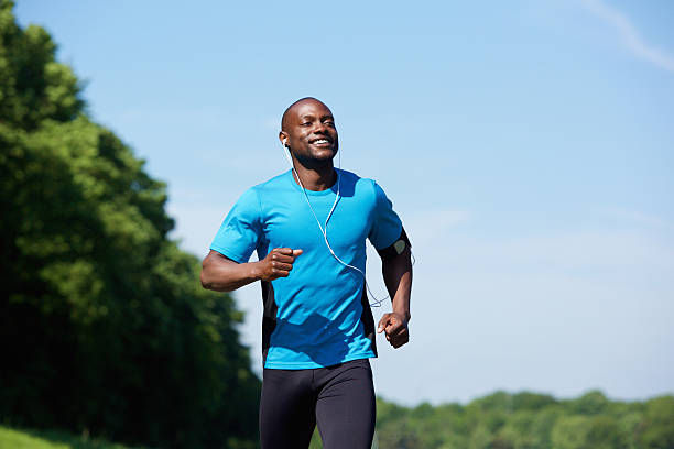 Man jogging outdoors, wearing blue shirt and earphones, looks happy. Background of trees and clear sky.