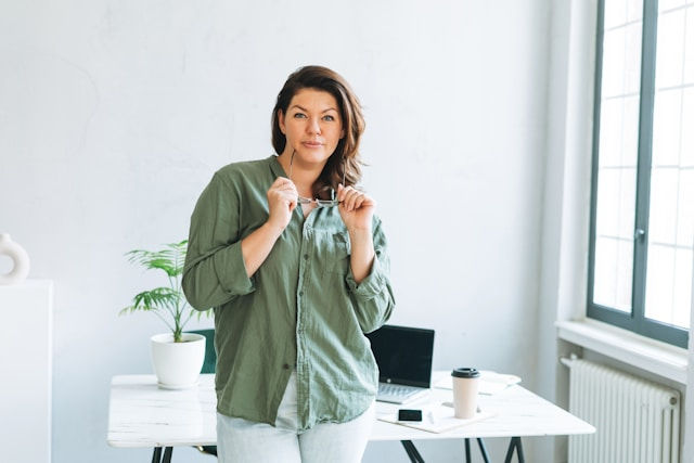 A confident woman stands in a bright, modern office holding her glasses, with a laptop, coffee cup, and plant on the desk behind her.