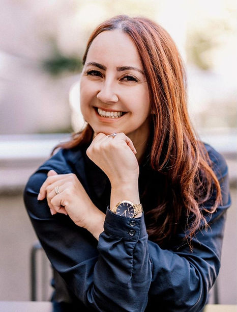 Smiling woman with long auburn hair, wearing a blue shirt and gold watch, rests chin on hand. 