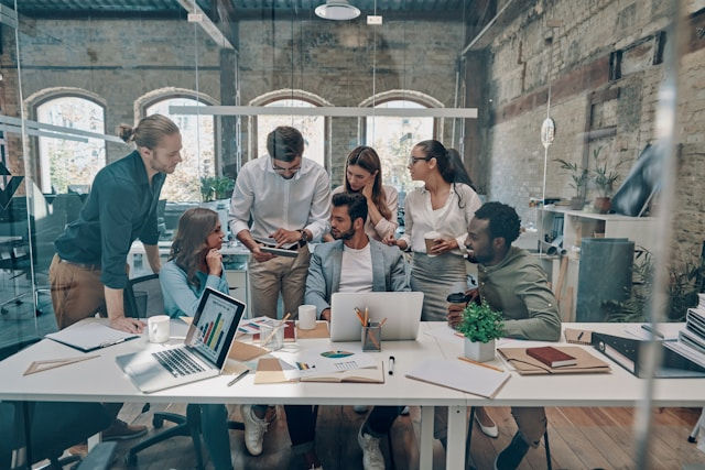 Team of coworkers huddles around a laptop in a bright office, reviewing charts and papers in a focused, collaborative meeting.