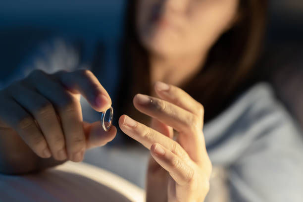 A woman is sitting in dim light, holding a ring and appearing to contemplate removing it from her finger, suggesting a moment of emotional reflection or decision.