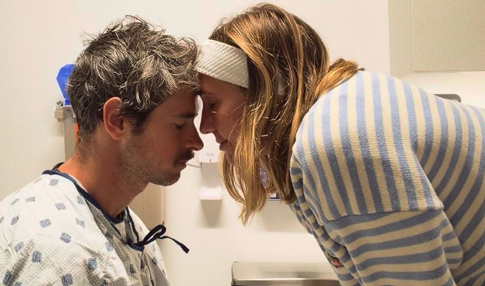 Couple touches foreheads in a hospital room; woman in striped sweater and headband comforts man in patient gown.