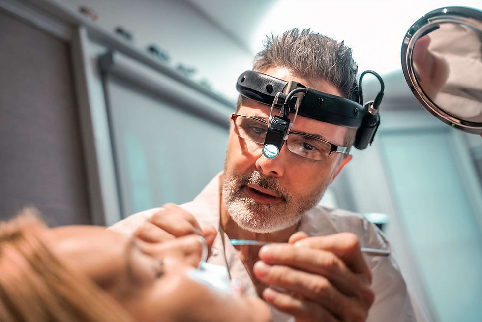 Dentist with headlamp and glasses examines patient's teeth with tools in a clinical setting. Focused expression, bright lighting.