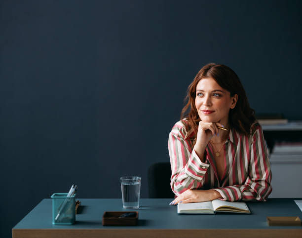 Woman in striped shirt pensively looks sideways, seated at a desk with a notebook and a glass of water, against a dark blue wall.