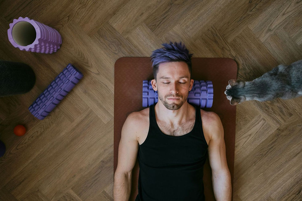 Man with purple hair lying on yoga mat using purple foam roller on neck. Cat approaches on wooden floor with various rollers nearby.