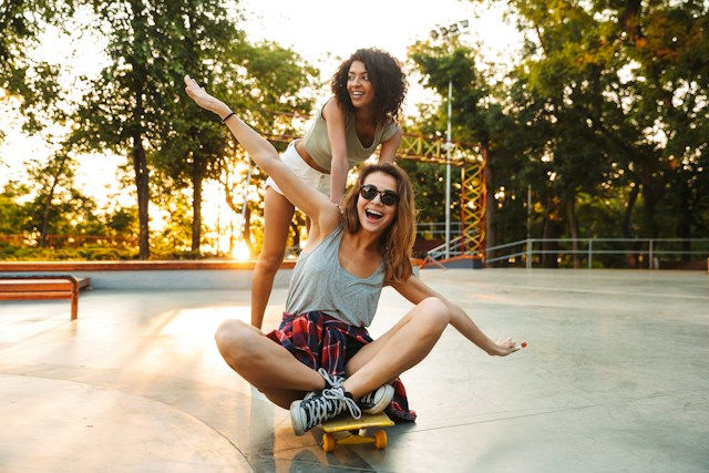 Two smiling women play on a skateboard at sunset in a park, one crouched in front and one balancing behind.