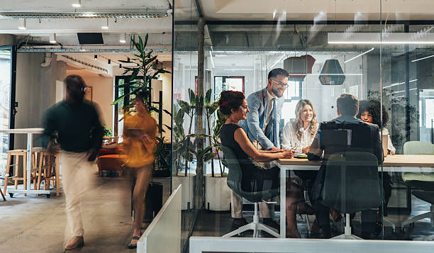 Meeting in a modern office with six people around a table, smiling and discussing. Motion blur of two people walking outside the glass room.