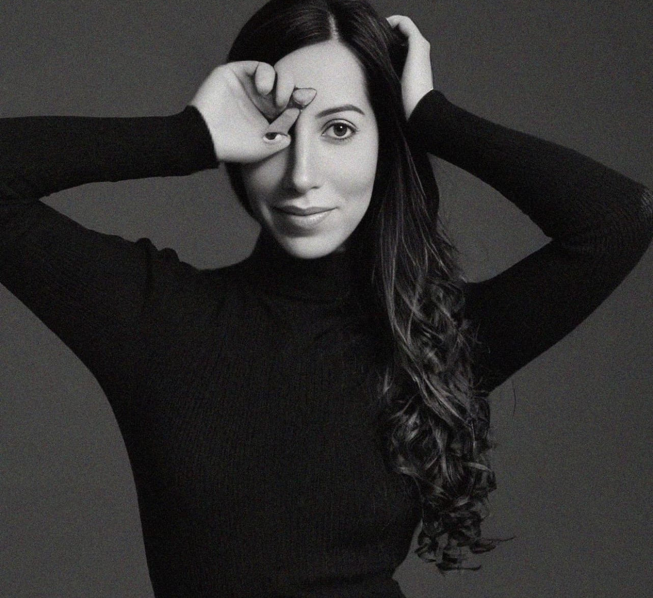 Black-and-white portrait of a woman in a black turtleneck, hands framing her face against a dark studio background.