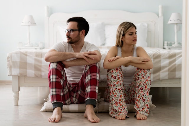 A couple sits back to back on the floor beside a bed, looking away from each other with tense expressions, suggesting conflict or emotional distance in a relationship.
