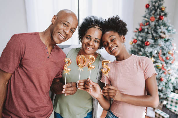 The photo shows a smiling family of three—father, mother, and daughter—standing together in front of a decorated Christmas tree, holding gold balloons that spell out "2025." 