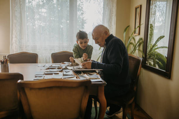 Elderly man and young boy look at photos on a wooden table. Cozy room with a window, plants, and chairs. Warm and nostalgic atmosphere.