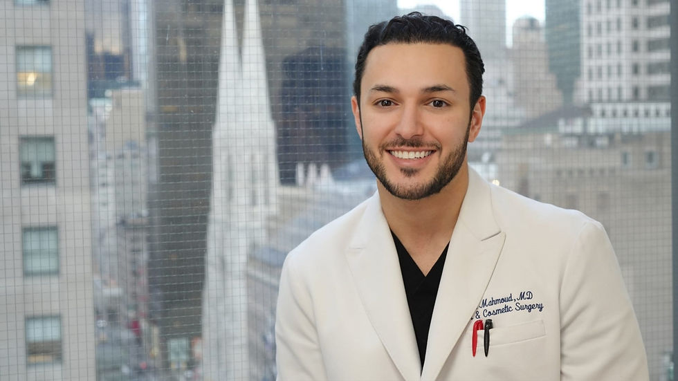 Smiling man in a white doctor’s coat stands by a window with a city skyline; coat reads Mahmoud, MD & Cosmetic Surgery.