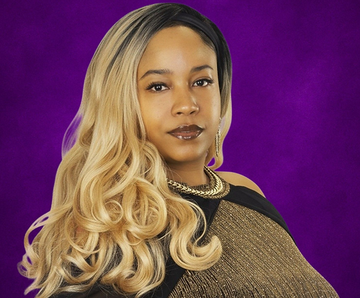 Portrait of a woman with long blonde curls in a gold-and-black dress against a purple background, gazing calmly at the camera.