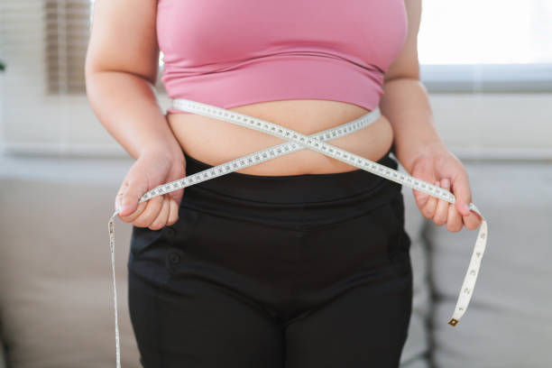 Person in a pink top and black pants measures their waist with a tape measure, standing in a bright room with a neutral background.
