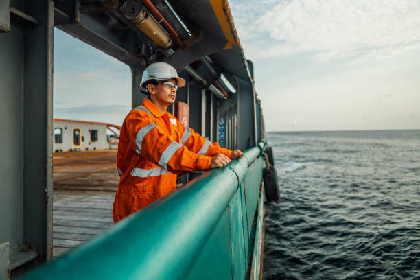 Worker in orange safety gear and helmet gazes at the ocean from a ship deck. Calm demeanor, clear sky, and vast sea background.