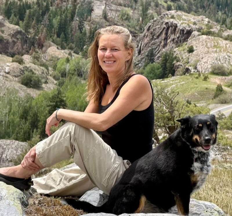 Smiling woman in a black tank top sits on a rock beside a black dog in a rocky mountain landscape.