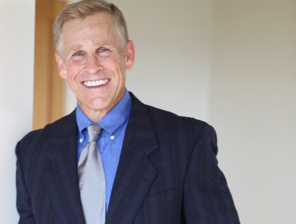 Smiling man in a suit, blue shirt, and gray tie stands indoors against a light-colored wall, conveying a cheerful mood.