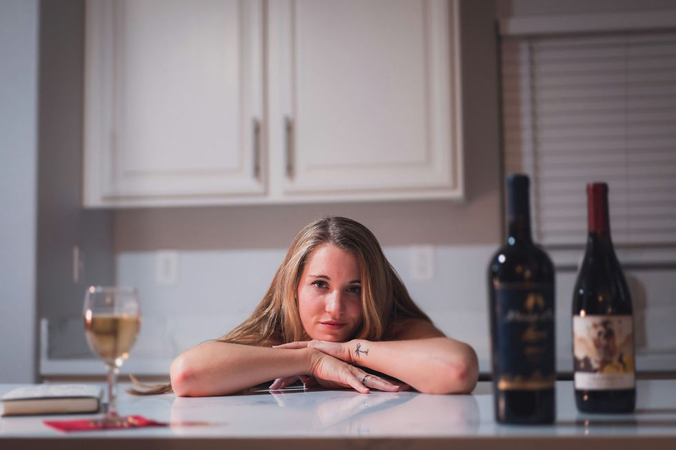 A woman with a tattoo leans on a counter, surrounded by wine bottles and a glass. She's in a kitchen, appearing relaxed and contemplative.
