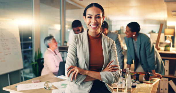 Smiling woman in a blazer stands with arms crossed in a bright office meeting, coworkers collaborating behind her.