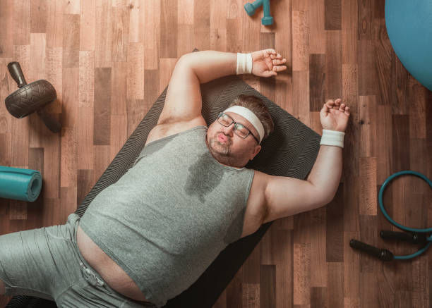 The image shows a tired man lying on a workout mat, wearing a gray tank top, headband, and wristbands.