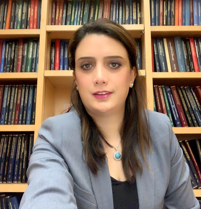 A person in a gray blazer sits in front of bookshelves filled with colorful books, wearing a turquoise necklace. Their expression is neutral.