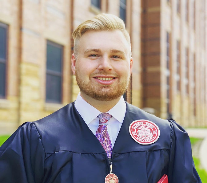 Smiling graduate in cap and gown with "Illinois State University" badge, standing near a brick building.