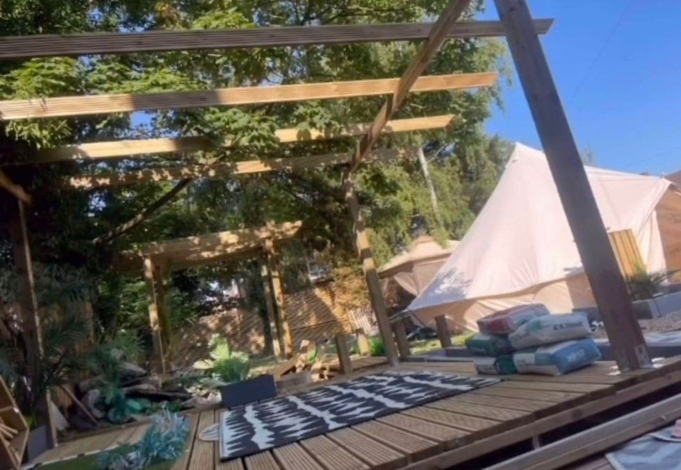 Wooden deck under construction with beams overhead in a lush garden. A tent and stacked cement bags are visible. Sunlight creates shadows.