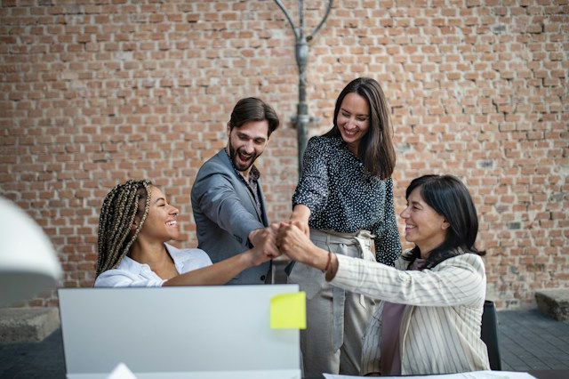 Four coworkers stack hands in a team huddle by a laptop, smiling in front of a brick wall.