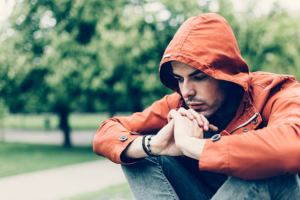 Man in orange hoodie sits pensively in a park, hands clasped, against a blurred leafy background, conveying a contemplative mood.