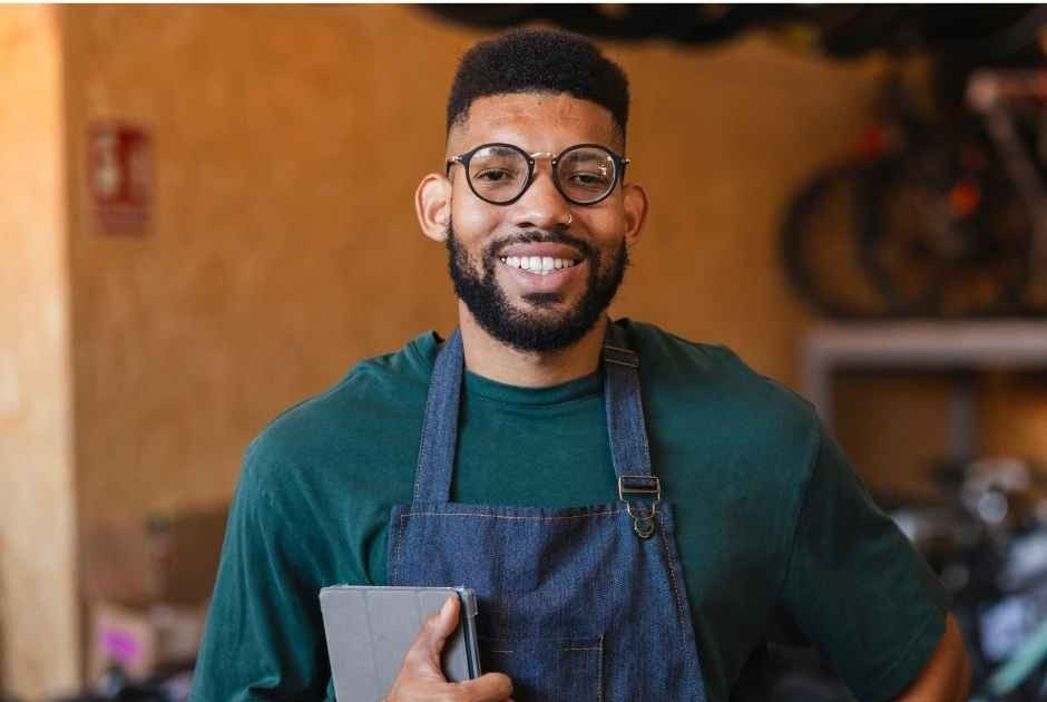 Smiling man with glasses and apron holds a tablet in a warm café or workshop setting.
