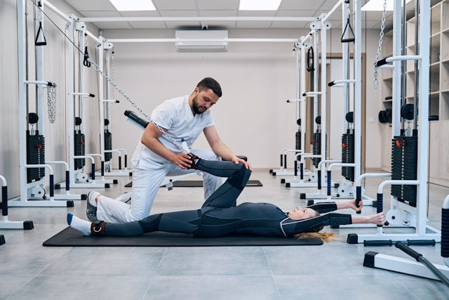 Therapist assists a woman in stretching on a mat in a bright gym, surrounded by exercise equipment. The mood is focused and supportive.