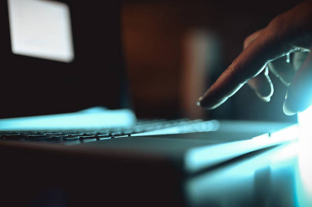 Hand hovers over a laptop keyboard in a dark room, lit by blue glow, creating a moody tech scene.