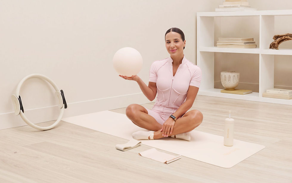 Woman in pink workout attire sits on a mat holding a ball in a minimal room with light wood floor and white shelves. Relaxed mood.