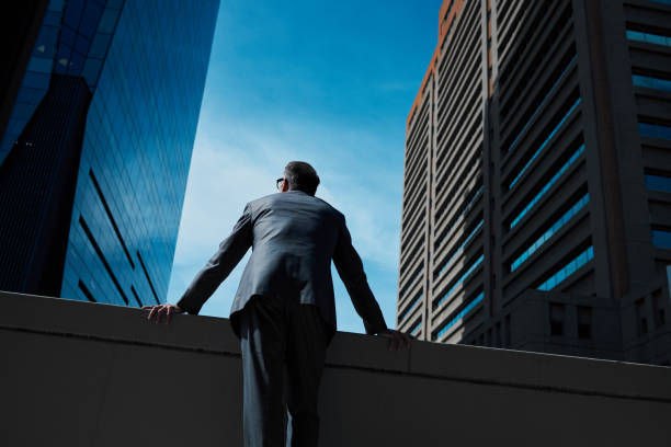 A man in a suit stands on a rooftop, gazing out at tall city buildings under a clear blue sky.