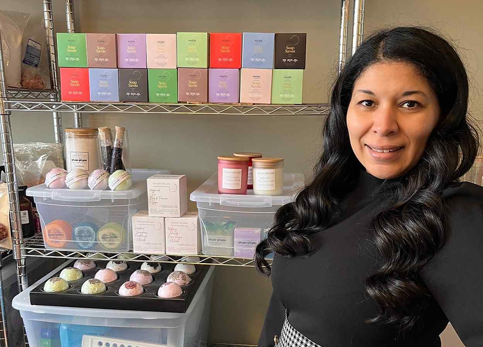 Woman in black top and checkered skirt smiling, standing by shelves with colorful packages and candles in a room. Bright, organized setting.