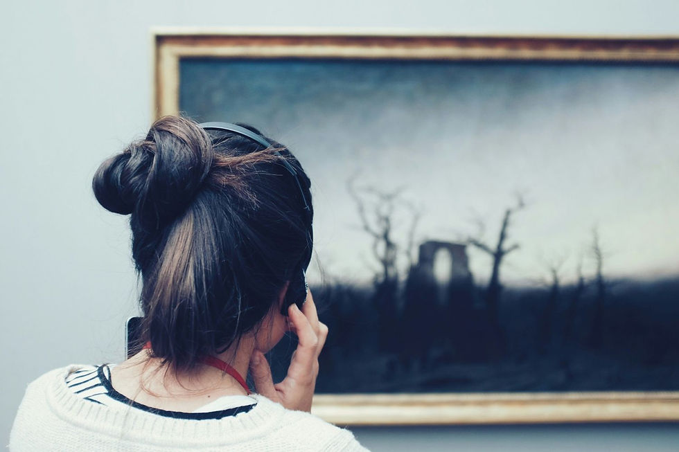 Woman with headphones viewing a dark, moody landscape painting in a gallery. She wears a white sweater, hair in bun, thoughtful mood.