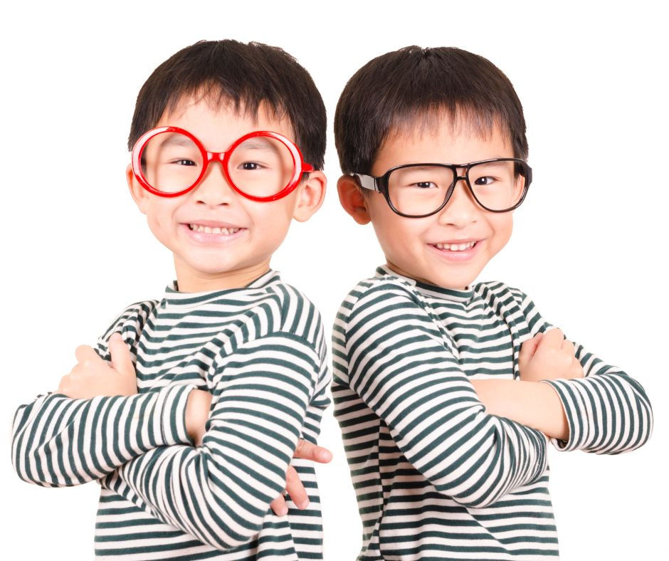 Two smiling twin boys wearing matching striped shirts and oversized glasses, one in red frames and the other in black frames, with their arms crossed confidently.