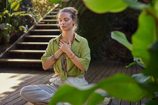 Woman meditating cross-legged on a wooden deck, hands on chest, with green foliage and sunlit stairs behind her.