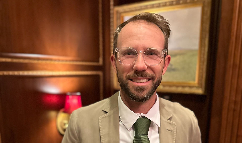 Smiling man in glasses, beige suit and green tie in a wood-paneled room with a framed painting and red lamp.