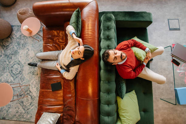 Overhead view of a man and woman relaxing on adjacent leather and green velvet sofas, holding pillows, in a cozy, modern living room setting.