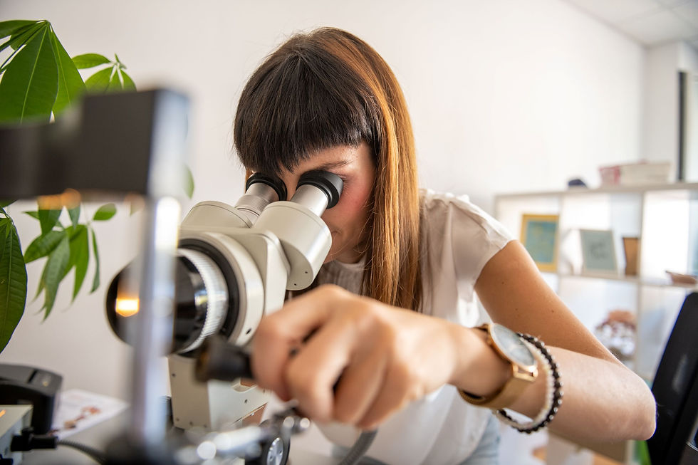 Woman peers into a microscope in a bright office lab, focused as she adjusts it; green plant and shelves blur in background