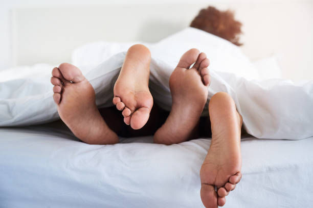 Two pairs of bare feet sticking out from under a white duvet, lying on a bed. Cozy and relaxed morning setting with soft lighting.