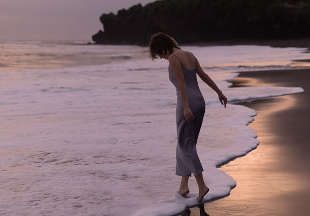 A woman in a long dress walks barefoot along the shoreline at sunset, gently touching the incoming waves.