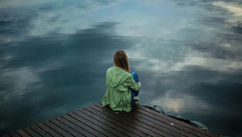 A person in a green jacket sits on a wooden dock, gazing at a calm, reflective lake under a cloudy sky, conveying solitude and peace.