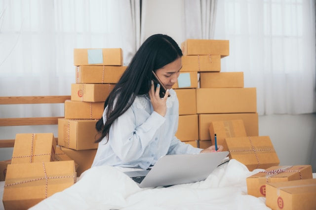 A young woman sitting on a bed surrounded by neatly stacked brown cardboard packages. She is talking on the phone while writing in a notebook and using a laptop, suggesting she is managing an online business or handling orders.